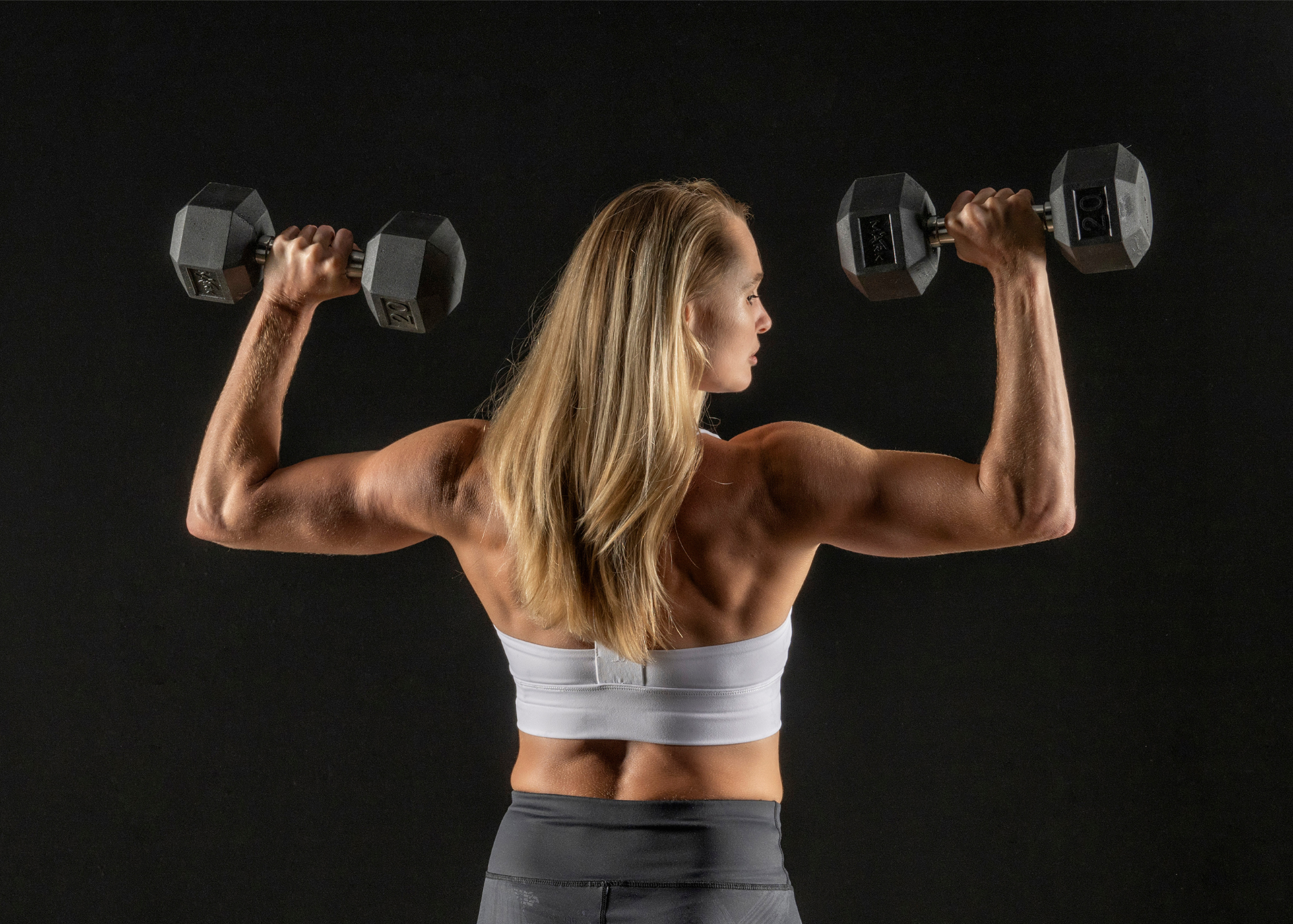 Female facing facing backward doing overhead press with dumbbells.