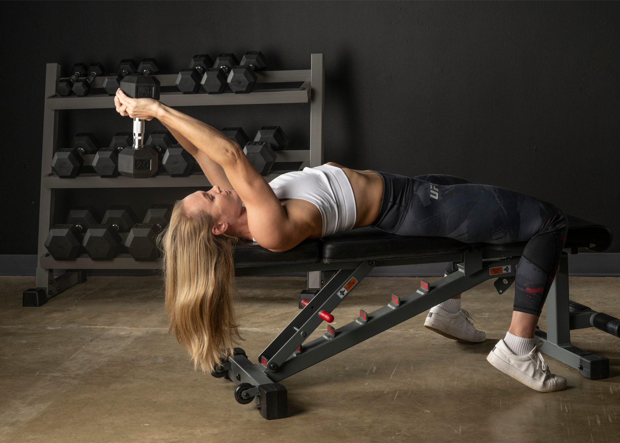 Female laying on flat bench doing skull crusher exercise with dumbbell over her head.