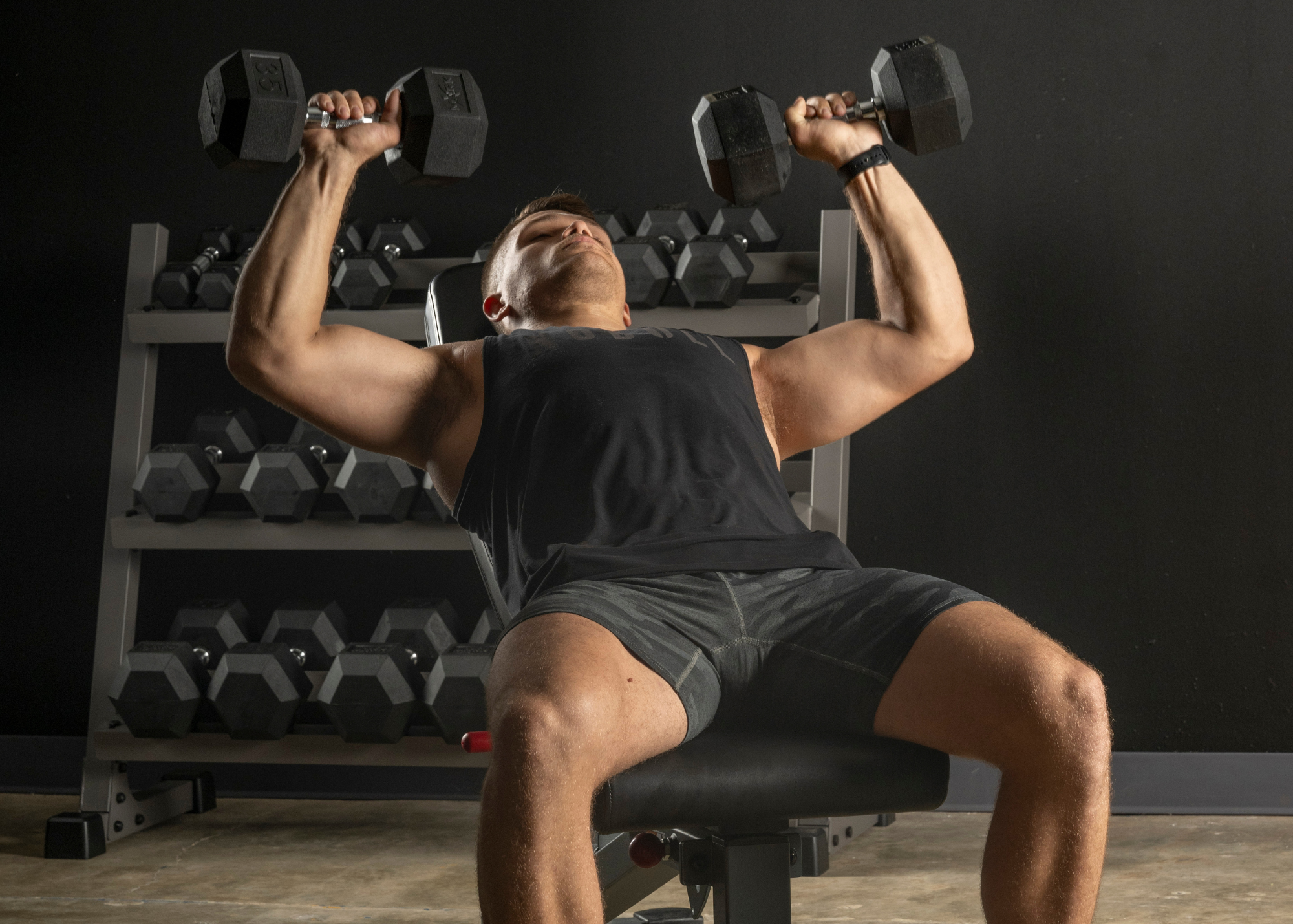 Male in reclined position on bench doing bench press with hex dumbbells.