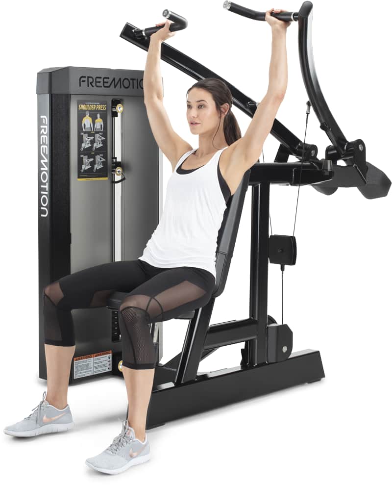 A woman in a white tank top and black leggings sits on a black Freemotion cable  machine, pushing the handles overhead. 