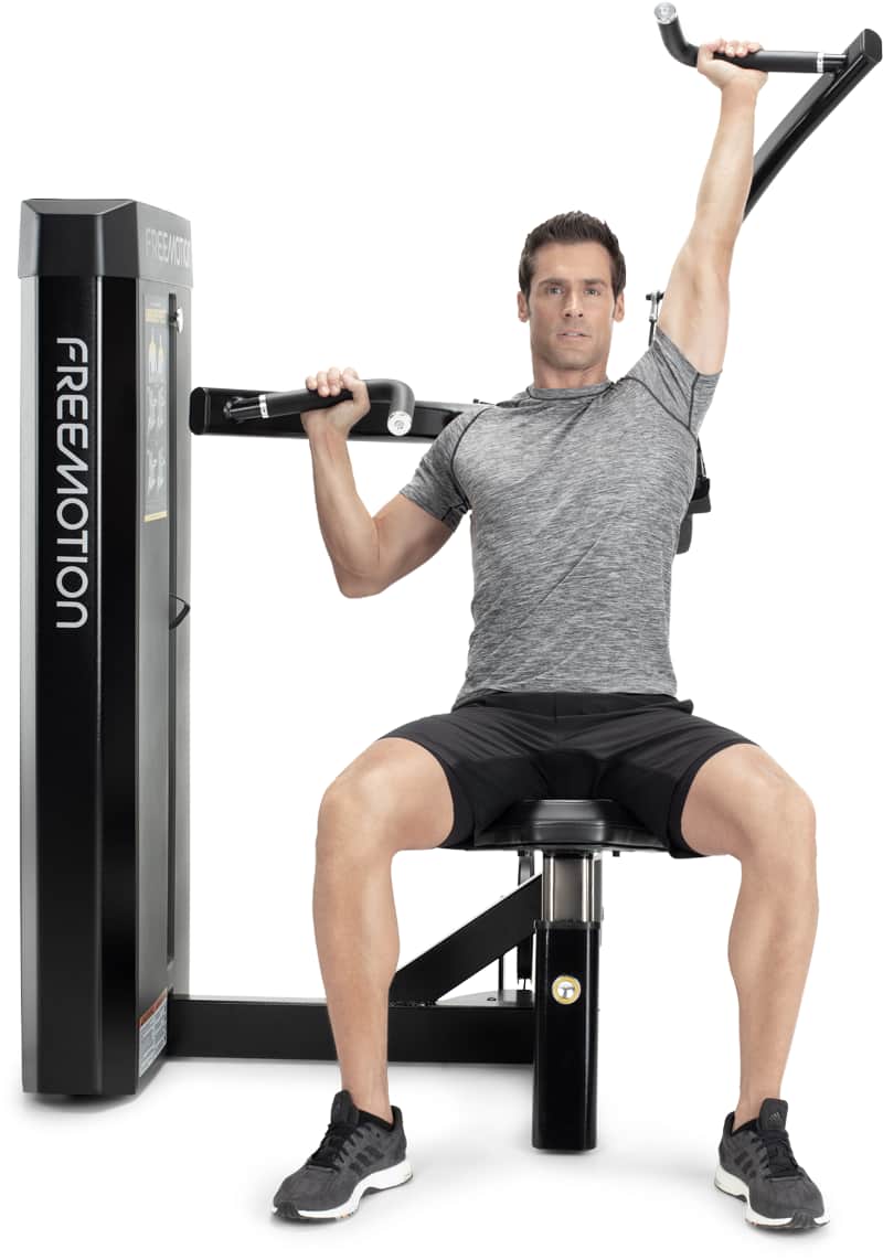 A man demonstrates the use of a Freemotion chest press exercise machine, seated on the bench with one arm overhead and the other resting on the bar, against a solid white background.