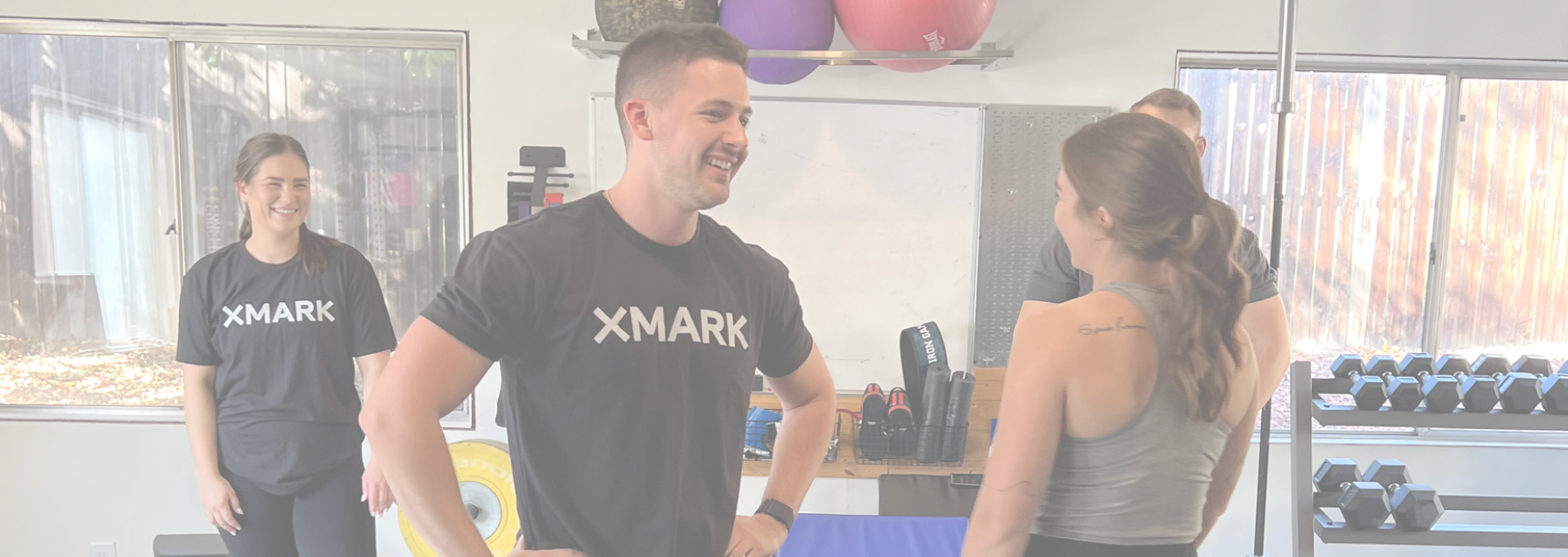 Three smiling people wearing XMARK tshirts in a gym